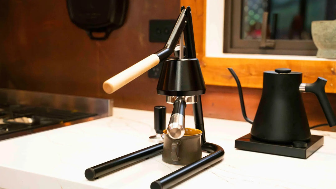 Manual espresso machine and coffee cup on marble bench top next to kettle.